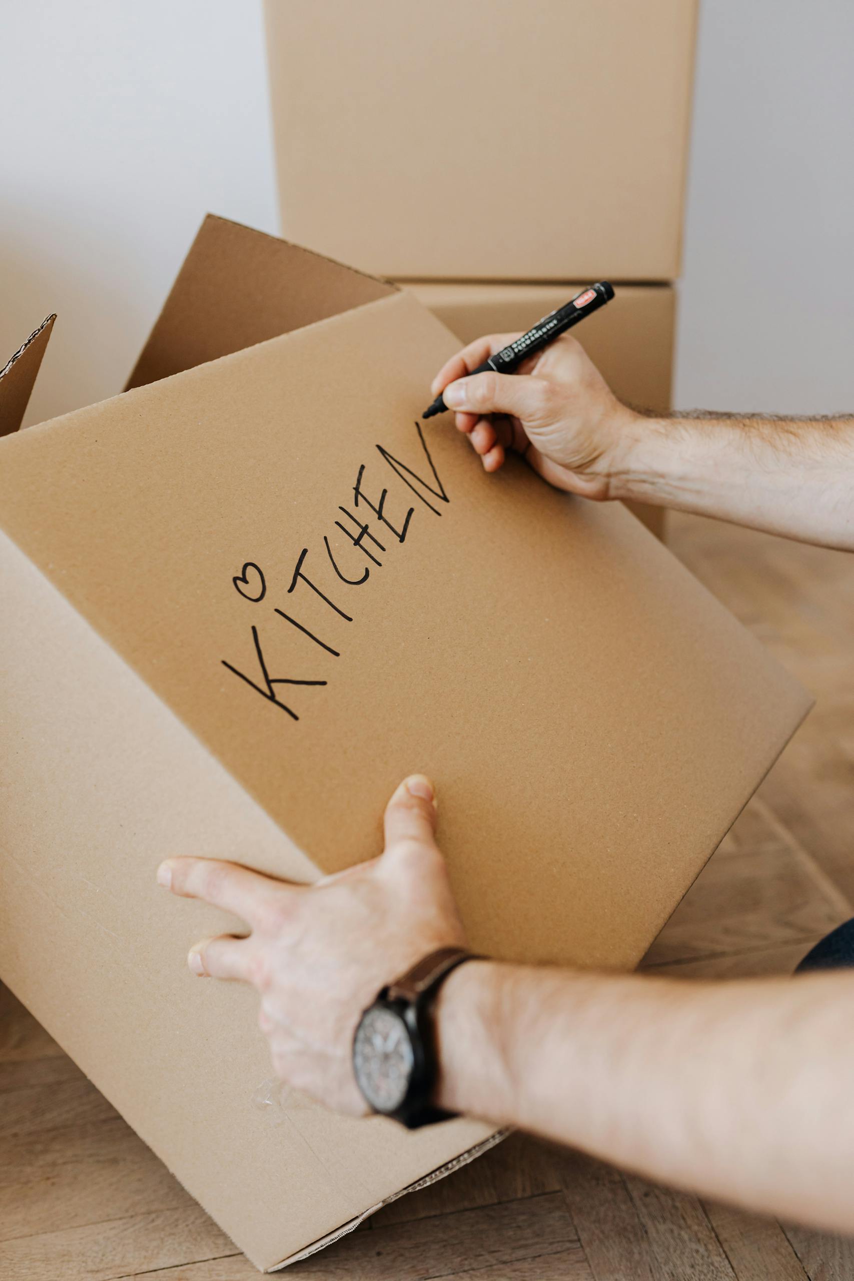 A man labels a cardboard box 'kitchen' during a home moving process. Indoors setting.